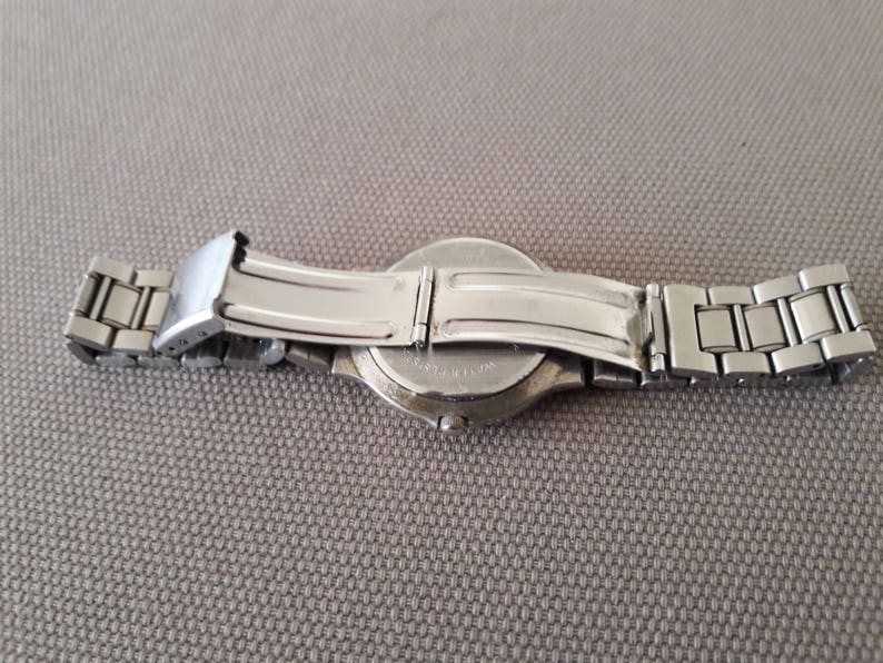 Puede incluir: Un reloj de pulsera plateado con una correa de acero inoxidable. El reloj est&aacute; sobre una superficie gris con la correa doblada hacia atr&aacute;s.