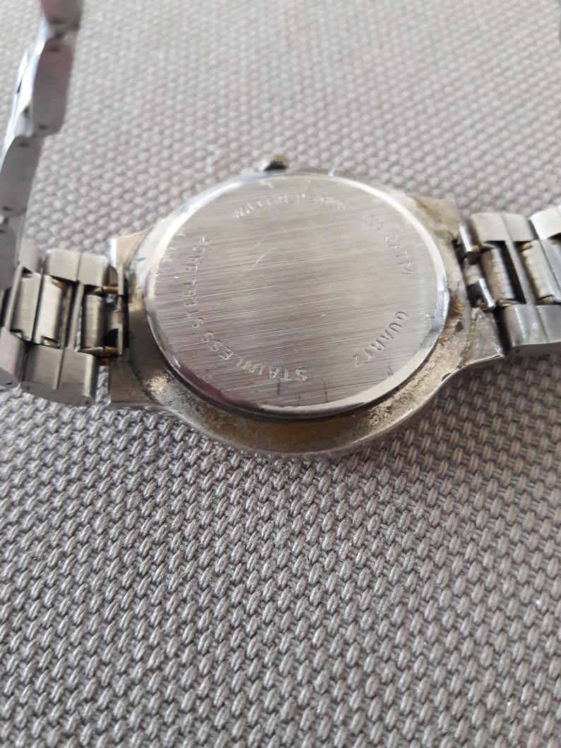 Puede incluir: La parte posterior de un reloj de pulsera plateado con una correa de acero inoxidable. La esfera del reloj est&aacute; en blanco y la parte posterior del reloj tiene el texto "WATER RESISTANT", "STAINLESS BACK", "22MM", "QUARTZ" y "JAPAN MVT".