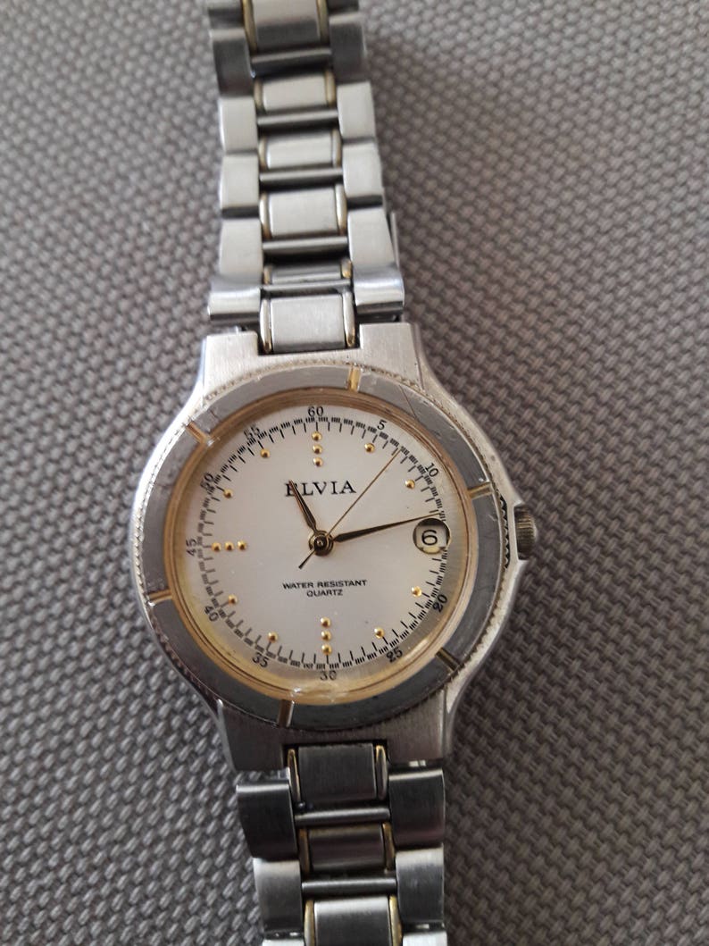 Puede incluir: Un reloj de pulsera de tono plateado y dorado con una esfera blanca y agujas negras. La esfera del reloj tiene las palabras "Relvia Water Resistant Quartz" impresas.