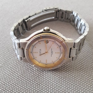 Puede incluir: Un reloj de pulsera plateado y dorado con una esfera blanca y agujas negras. La esfera del reloj tiene las palabras "Ilvia" y "Water Resistant Quartz" impresas.