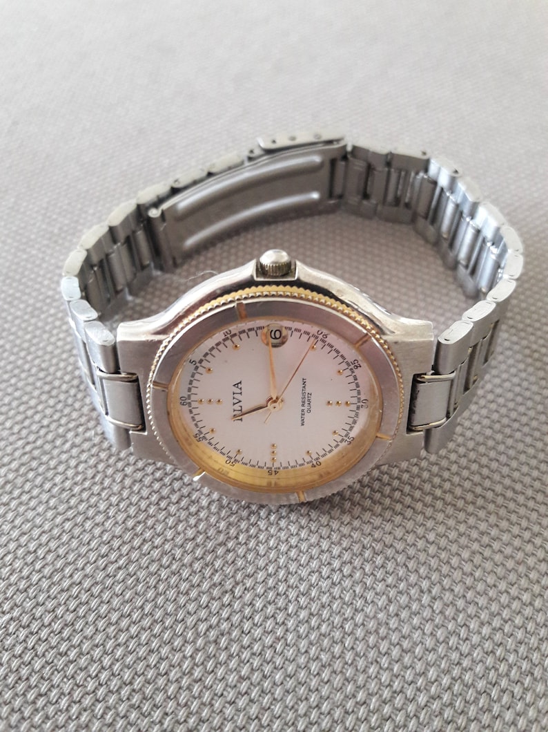 Puede incluir: Un reloj de pulsera plateado y dorado con una esfera blanca y agujas negras. La esfera del reloj tiene las palabras "Ilvia" y "Water Resistant Quartz" impresas.