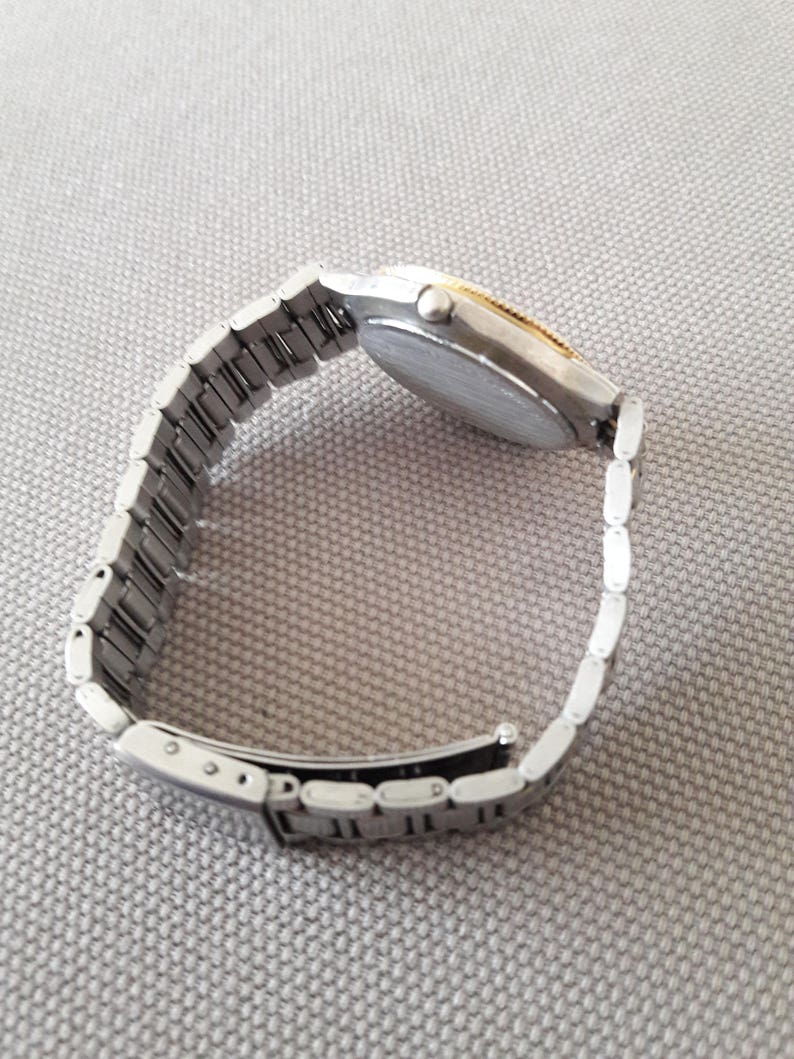 Puede incluir: Un reloj de pulsera plateado y dorado con una banda de metal plateado. El reloj tiene una esfera redonda y un bisel plateado.