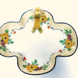 Op de afbeelding: Een witte porseleinen serveerschaal met een gouden handvat en een bloemenmotief. De schaal heeft een gegolfde rand en is versierd met gele, oranje en paarse bloemen.
