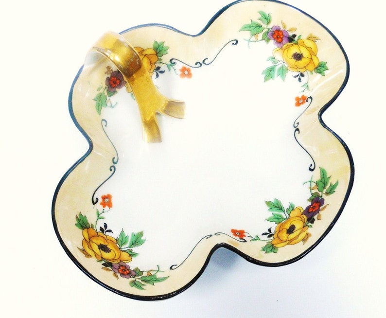 Op de afbeelding: Een witte porseleinen serveerschaal met een gouden handvat en een bloemenmotief. De schaal heeft een gegolfde rand en is versierd met gele, oranje en paarse bloemen.