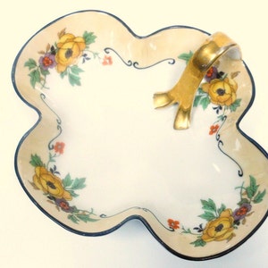 Op de afbeelding: Een witte porseleinen serveerschaal met een gouden handvat en een bloemenmotief. De schaal heeft een gegolfde rand en is versierd met gele, oranje en paarse bloemen.