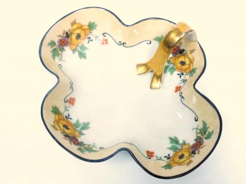 Op de afbeelding: Een witte porseleinen serveerschaal met een gouden handvat en een bloemenmotief. De schaal heeft een gegolfde rand en is versierd met gele, oranje en paarse bloemen.