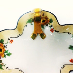 Op de afbeelding: Een witte porseleinen schaal met een gouden handvat en een bloemenmotief. De schaal heeft een gegolfde rand en is versierd met gele, oranje en paarse bloemen.