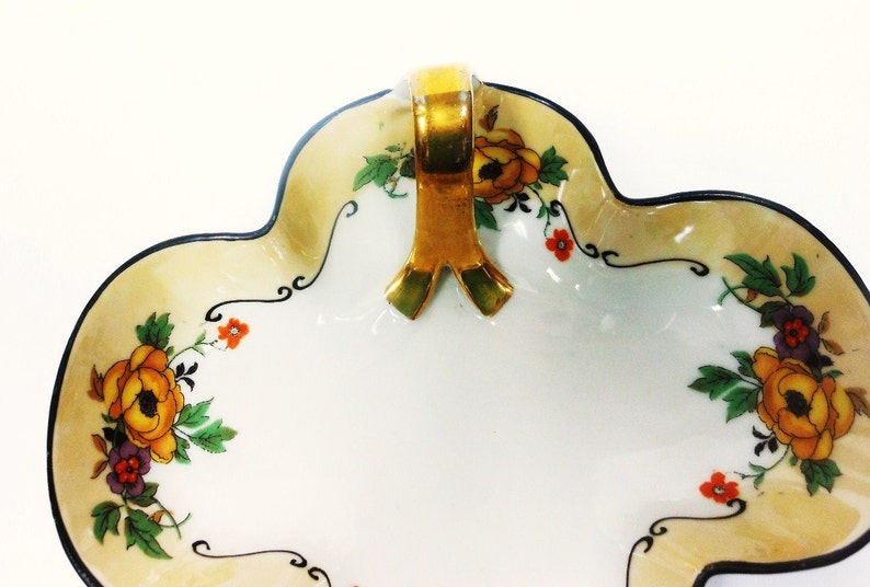 Op de afbeelding: Een witte porseleinen schaal met een gouden handvat en een bloemenmotief. De schaal heeft een gegolfde rand en is versierd met gele, oranje en paarse bloemen.