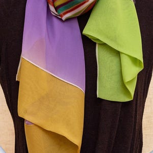May include: A colorful silk scarf with a vibrant striped pattern and solid panels of purple, yellow, and green. The scarf is draped on a mannequin, showcasing its flowing design and the text "ATELIER MAISON DE SOIE".