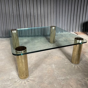 May include: A square glass coffee table with four gold metal cylindrical legs. The table has a clear glass top and a polished gold finish on the legs.