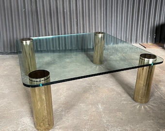 May include: A square glass coffee table with four gold metal cylindrical legs. The table has a clear glass top and a polished gold finish on the legs.