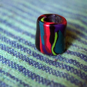 Könnte beinhalten: Eine bunte, mehrfarbige, zylindrische Perle mit einem Loch in der Mitte. Die Perle ist aus Polymer Clay gefertigt und hat ein Farbschema in Schwarz, Rot, Grün und Lila.
