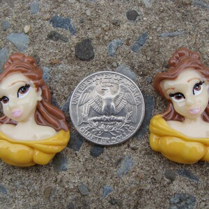 May include: Two yellow and brown Belle character figurines from the Disney movie Beauty and the Beast. The figurines are about the size of a US quarter.