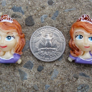May include: Two purple and white princess-themed buttons with brown hair and crowns. The buttons are about the size of a US quarter.