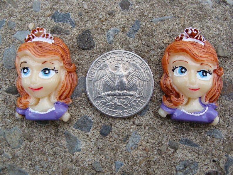 May include: Two purple and white princess-themed buttons with brown hair and crowns. The buttons are about the size of a US quarter.