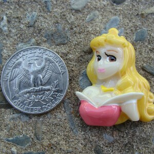May include: A yellow and pink plastic figurine of a princess with long blonde hair. She is wearing a pink dress and a gold necklace.