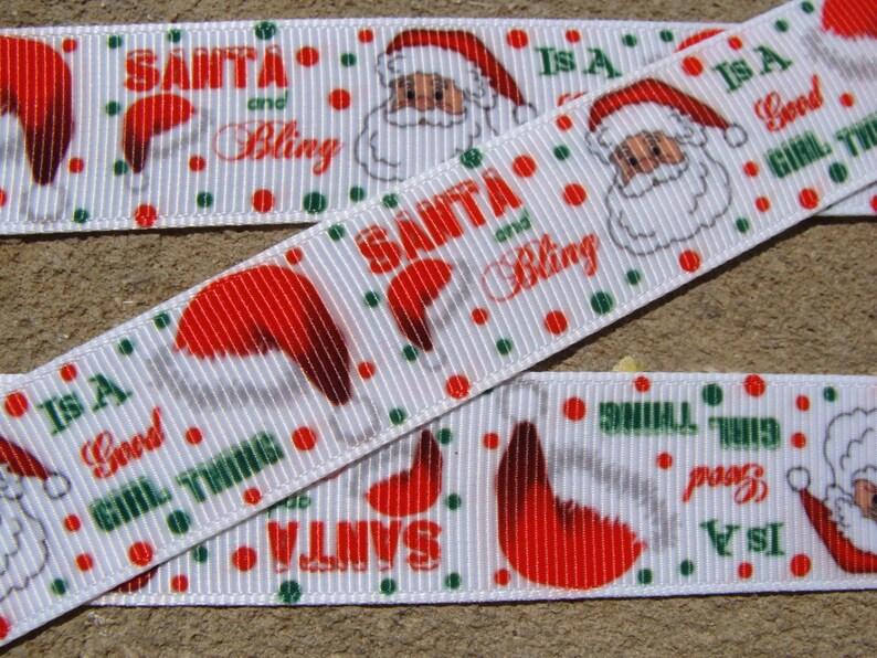 May include: White ribbon with a repeating pattern of Santa Claus, a red hat, and the text "Santa and Bling" and "Is a Good Girl Thing".