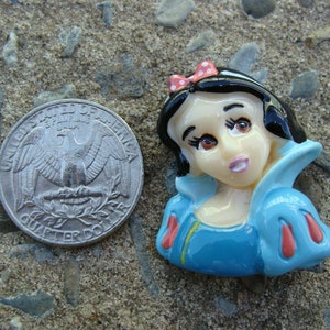 May include: A porcelain figurine of Snow White, a Disney princess, wearing a blue dress with red accents and a red bow in her black hair.