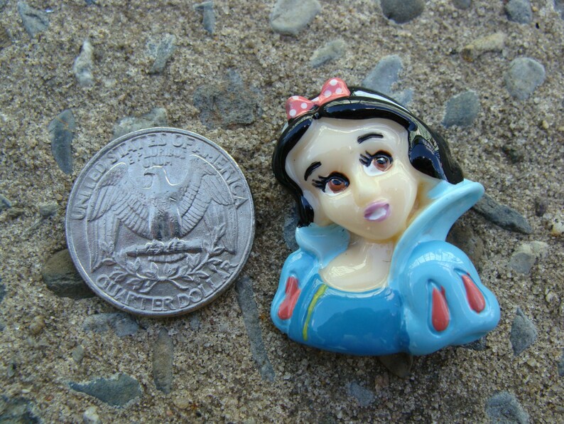 May include: A porcelain figurine of Snow White, a Disney princess, wearing a blue dress with red accents and a red bow in her black hair.