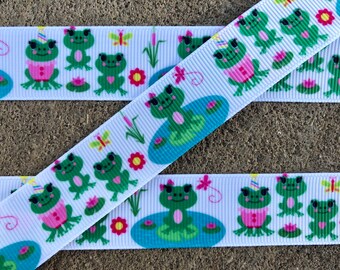 May include: White ribbon with a repeating pattern of green frogs wearing party hats, butterflies, flowers, and lily pads. The frogs are wearing pink shirts and have green eyes.