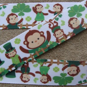 May include: White ribbon with a repeating pattern of green shamrocks, brown monkeys wearing green shirts, and some monkeys wearing green hats. The monkeys are holding onto a white fence.