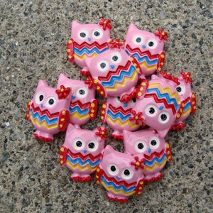 May include: A collection of nine pink owl buttons with colorful zig-zag patterns and yellow accents. The owls have black eyes and are decorated with red and yellow flowers.