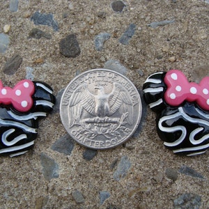 May include: Two black and white ceramic Minnie Mouse head ornaments with pink polka dot bows. The ornaments are about the size of a US quarter.