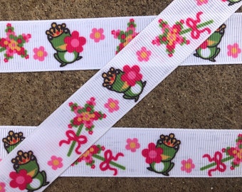 May include: White ribbon with a repeating pattern of green frogs wearing crowns, pink flowers, and green stars with pink and yellow centers.