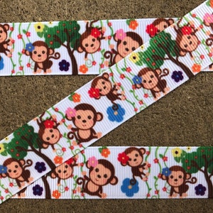 May include: A white grosgrain ribbon featuring a repeating pattern of brown cartoon monkeys, green trees, and colorful flowers in shades of red, pink, blue, and orange. The monkeys are depicted with smiling faces and curled tails among vines.
