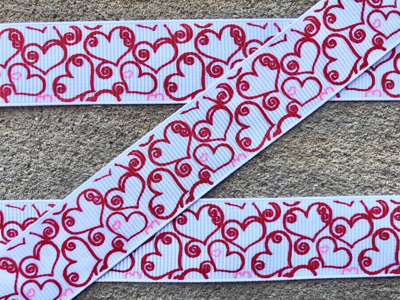 May include: White ribbon featuring a repeating pattern of red hearts and swirls. The hearts have a decorative design against a light blue striped background. Small pink hearts are also present.