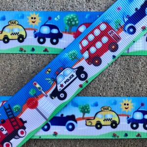 May include: A blue and white ribbon with a repeating pattern of various vehicles, including a red double-decker bus, a yellow taxi, a blue car, a red fire truck, a blue police car, and a blue truck. The ribbon has a white background with green grass and trees.