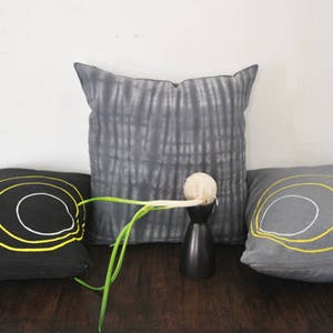 May include: Three decorative throw pillows with concentric circles in yellow and white on a black and gray background. The pillows are arranged on a dark wooden surface with a green plant and a white bulb in the center.