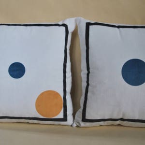 May include: Two white throw pillows with black borders and geometric designs. One pillow has a blue circle and an orange circle. The other pillow has a blue circle.