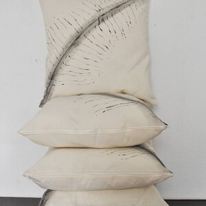 May include: A stack of four white cotton pillow covers with black and red abstract designs. The designs are hand-painted and resemble feathers.