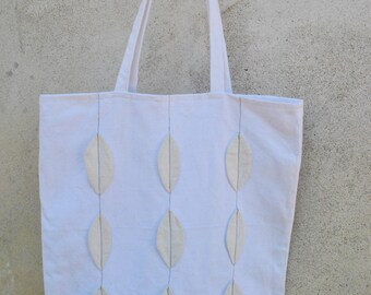 May include: A white canvas tote bag with a design of white leaf shapes sewn on. The bag has two long straps and is hanging from a wooden peg.