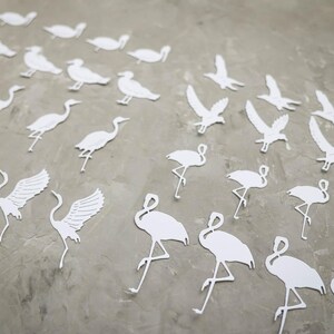 May include: White paper cutouts of various birds, including cranes, flamingos, and birds in flight, are scattered across a grey surface. The cutouts are detailed, with wings and legs visible, creating a decorative and artistic arrangement.