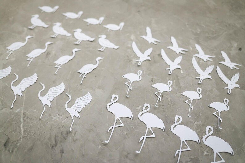 May include: White paper cutouts of various birds, including cranes, flamingos, and birds in flight, are scattered across a grey surface. The cutouts are detailed, with wings and legs visible, creating a decorative and artistic arrangement.