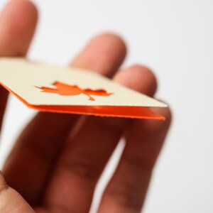 May include: A close-up of a cream-colored tag with an orange maple leaf cutout. The tag has an orange border and is held between fingers. The website mapleapple.eu is visible in the background.