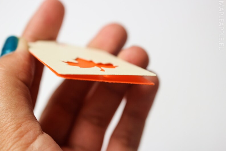 May include: A close-up of a cream-colored tag with an orange maple leaf cutout. The tag has an orange border and is held between fingers. The website mapleapple.eu is visible in the background.