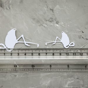 May include: Two white flamingo cutouts, each approximately 5 cm long, are positioned on a grey surface next to a clear ruler. The ruler shows measurements in both centimetres and inches. The cutouts are detailed and stylized.