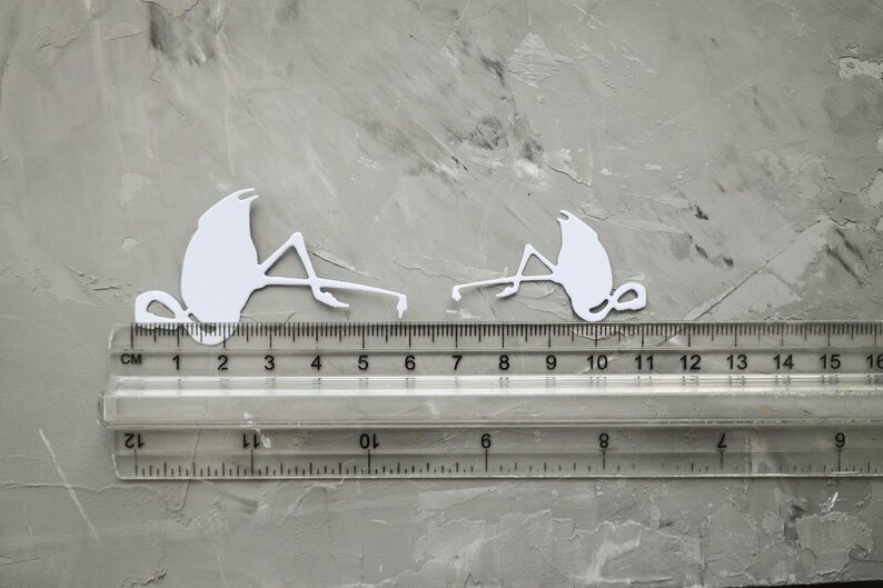 May include: Two white flamingo cutouts, each approximately 5 cm long, are positioned on a grey surface next to a clear ruler. The ruler shows measurements in both centimetres and inches. The cutouts are detailed and stylized.