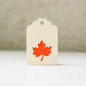 May include: A cream-colored tag with a scalloped top and a hole for hanging. The tag features a bright orange maple leaf design. The background is a neutral color, and the tag is on a white surface.