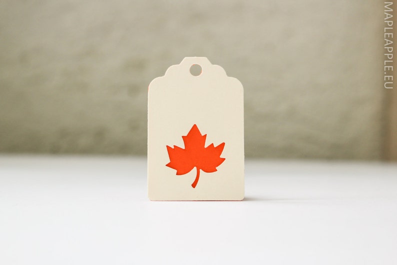 May include: A cream-colored tag with a scalloped top and a hole for hanging. The tag features a bright orange maple leaf design. The background is a neutral color, and the tag is on a white surface.
