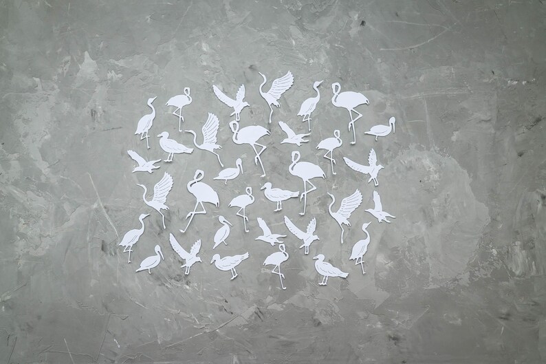 May include: A collection of white paper cutouts depicting various bird species, such as cranes and waterfowl, arranged on a grey, textured surface. The birds are shown in different poses, some with wings extended, others standing or in flight.