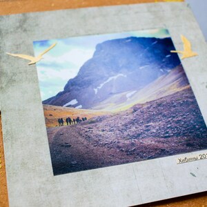 May include: A scrapbook page with a landscape photograph of a mountain range and hikers. The photo is framed with a grey border and decorated with two yellow bird cutouts. The page has a brown background with gold embellishments and a fabric tag.