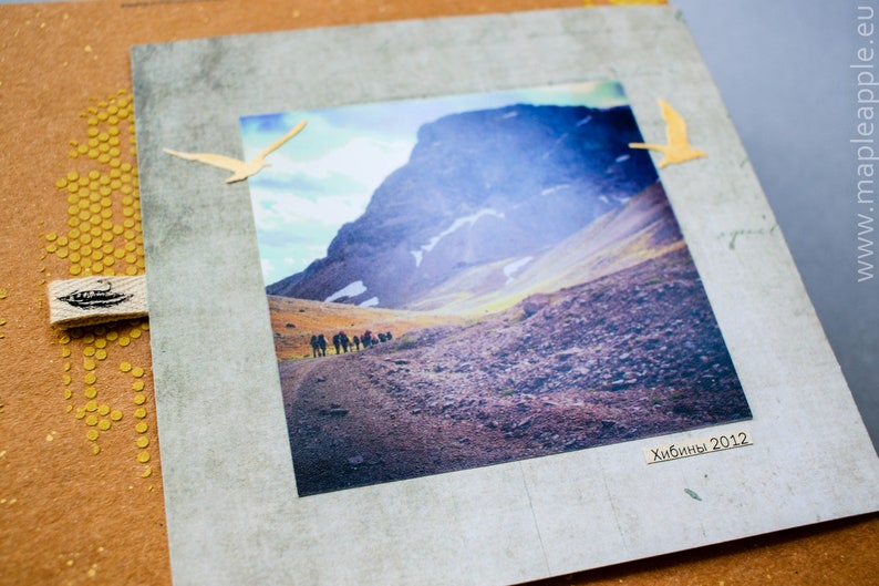 May include: A scrapbook page with a landscape photograph of a mountain range and hikers. The photo is framed with a grey border and decorated with two yellow bird cutouts. The page has a brown background with gold embellishments and a fabric tag.