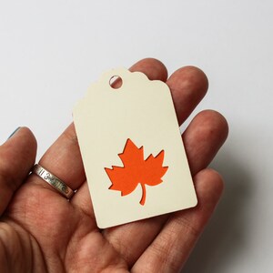 May include: A cream-colored tag with a die-cut orange maple leaf design. The tag has a scalloped top edge and a hole for hanging. The tag is held in a hand against a white background. The website mapleapple.eu is visible in the top right corner.