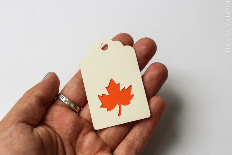 May include: A cream-colored tag with a die-cut orange maple leaf design. The tag has a scalloped top edge and a hole for hanging. The tag is held in a hand against a white background. The website mapleapple.eu is visible in the top right corner.