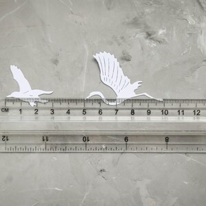 May include: Two white paper cutouts of birds in flight are positioned on a grey surface next to a clear ruler. The ruler shows measurements in centimetres. The birds are approximately 2 to 8 cm in length.