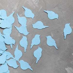 May include: Light blue bird-shaped cutouts scattered on a grey textured surface. The cutouts vary in shape, some with wings and others without, creating a whimsical and decorative effect. The image suggests a craft or DIY project.